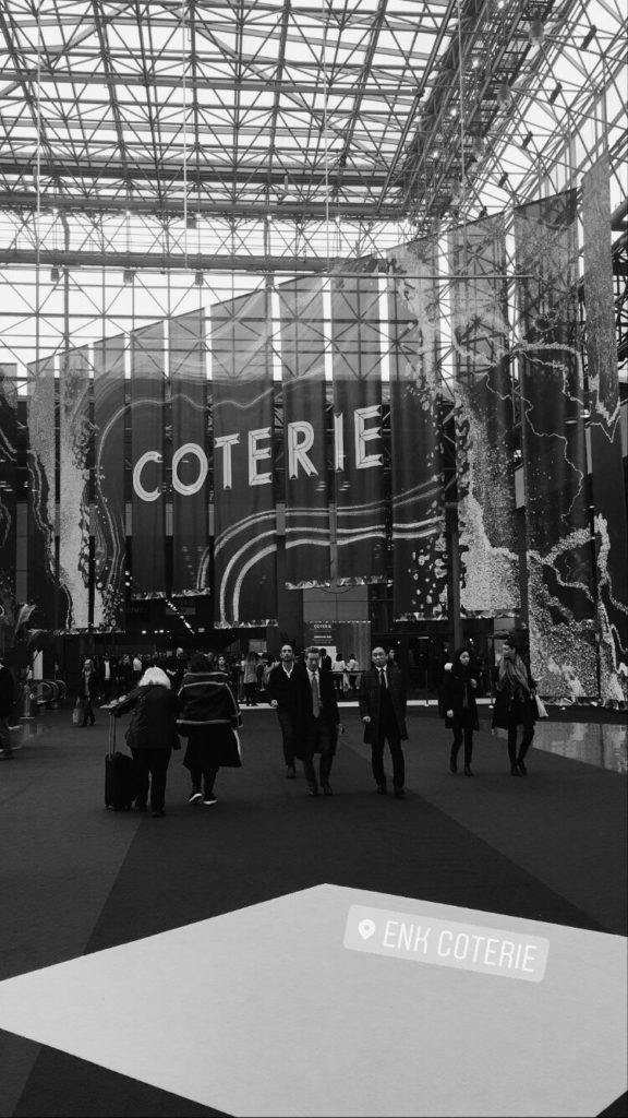 black and white photo of a group of people walking inside a convention center with the word coterie in block letters in the background